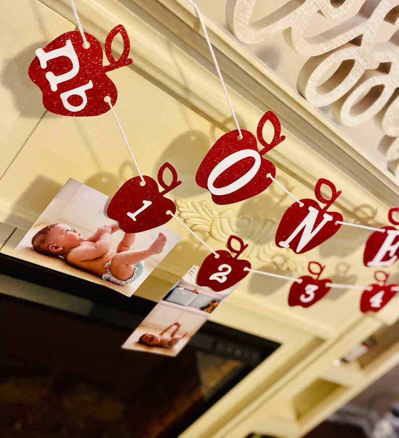 May include: Red glitter apple-shaped banner with white letters spelling "ONE" and numbers 1-4. The banner is strung with white string and has photos of a baby in each section.