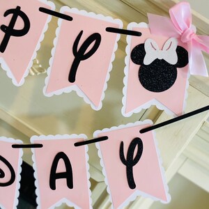 Puede incluir: Bandera rosa y negra con las palabras "Happy Day" y una silueta negra de Minnie Mouse con un lazo blanco. La bandera est&aacute; enhebrada con una cuerda negra y tiene bordes festoneados blancos.