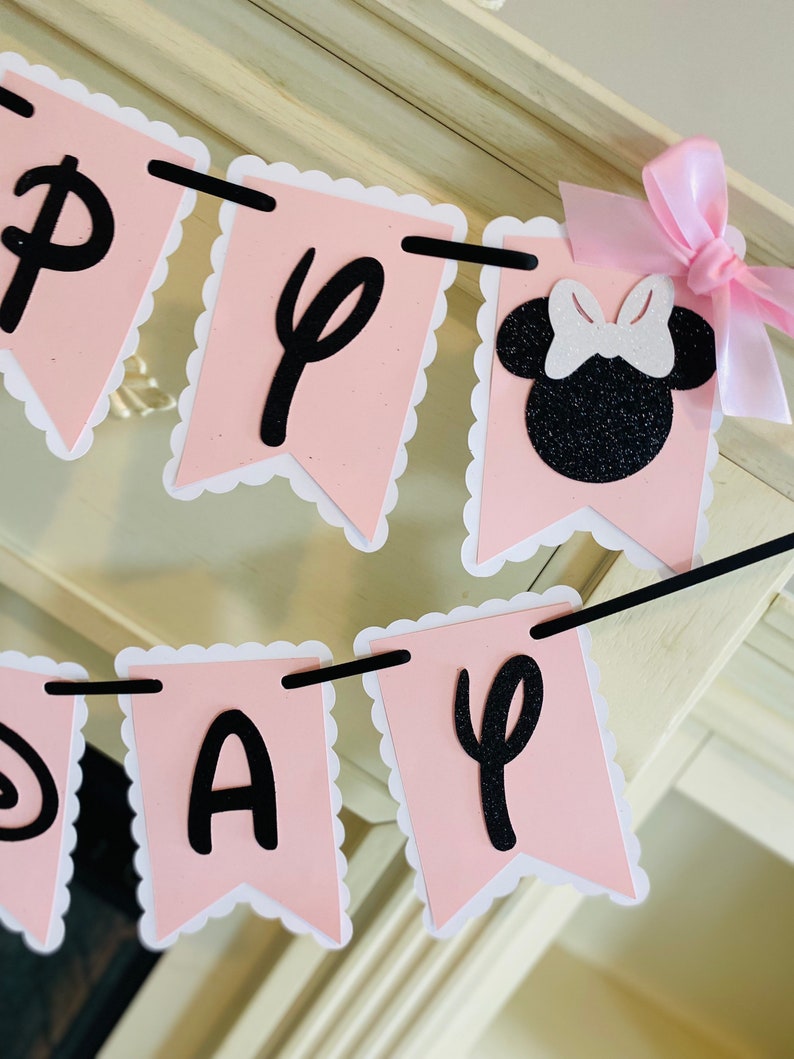 Puede incluir: Bandera rosa y negra con las palabras "Happy Day" y una silueta negra de Minnie Mouse con un lazo blanco. La bandera est&aacute; enhebrada con una cuerda negra y tiene bordes festoneados blancos.