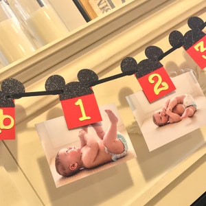 May include: A black and red Mickey Mouse themed banner with pictures of a baby. The banner has black Mickey Mouse ears with red and yellow numbers 1 and 2.