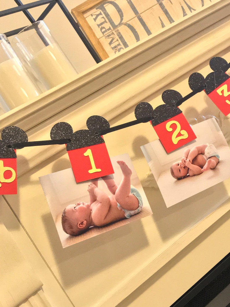 May include: A black and red Mickey Mouse themed banner with pictures of a baby. The banner has black Mickey Mouse ears with red and yellow numbers 1 and 2.