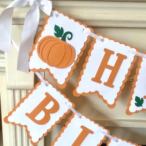 May include: A decorative banner featuring scalloped white paper panels with orange borders, strung together with white ribbon. The panels display orange letters spelling out a message and include cut-out orange pumpkin shapes with green glitter leaves.
