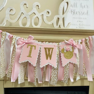 May include: A pink and gold "TW" banner with a Minnie Mouse silhouette, perfect for a second birthday celebration. The banner features pink and white ribbons, gold polka dot fabric, and gold glitter lettering. Decorative bows adorn the banner.