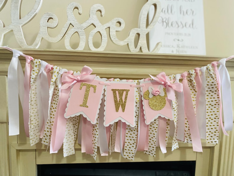 May include: A pink and gold "TW" banner with a Minnie Mouse silhouette, perfect for a second birthday celebration. The banner features pink and white ribbons, gold polka dot fabric, and gold glitter lettering. Decorative bows adorn the banner.