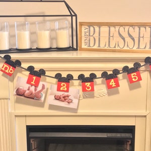 May include: A black and red Mickey Mouse themed baby milestone banner with photos of a baby boy. The banner has the numbers 1 through 5 and the word "nb" for newborn. The banner is hanging above a fireplace.