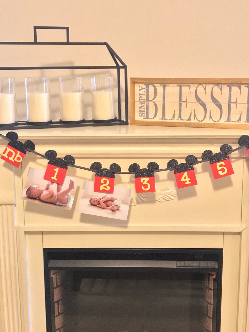 May include: A black and red Mickey Mouse themed baby milestone banner with photos of a baby boy. The banner has the numbers 1 through 5 and the word "nb" for newborn. The banner is hanging above a fireplace.