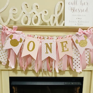 May include: A pink and gold "ONE" banner with Minnie Mouse silhouettes, polka dots, and ribbons. The banner is part of a first birthday party decoration. A sign with the text "Her Children Rise Up and call her Blessed" is in the background.