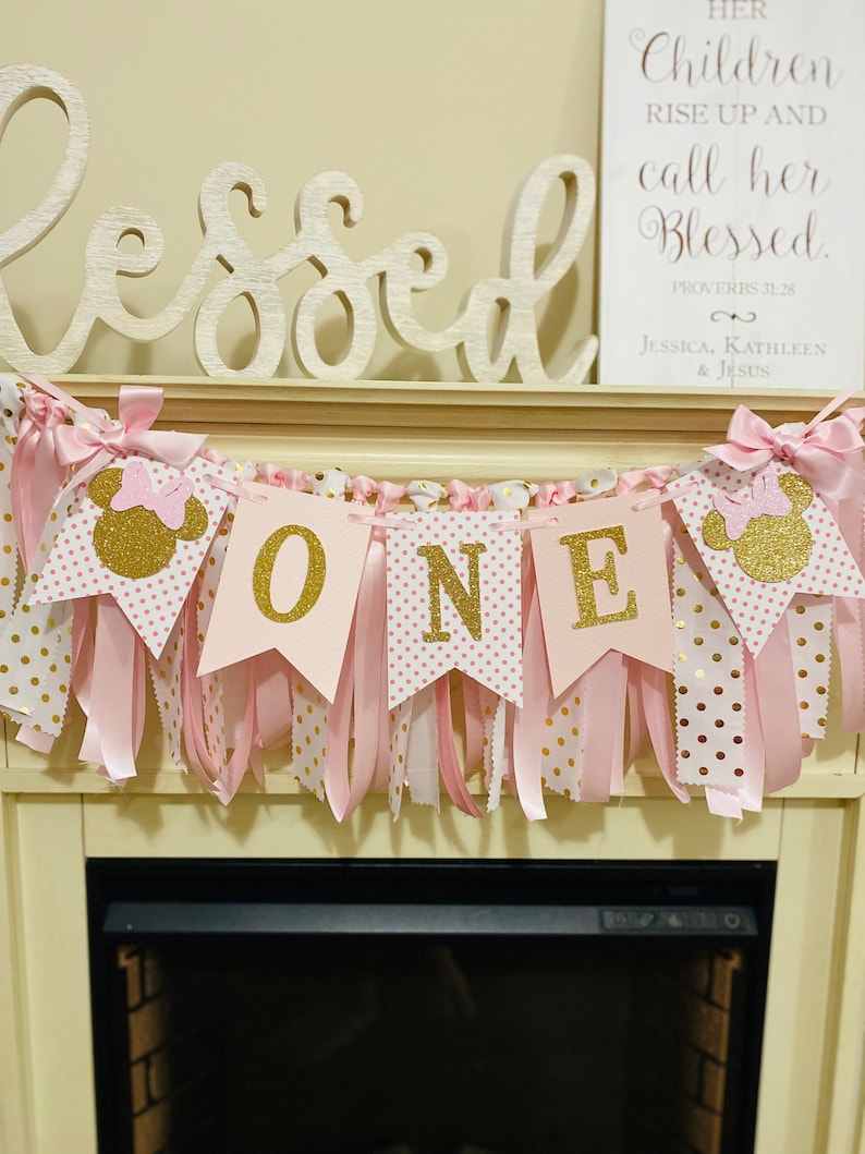 May include: A pink and gold "ONE" banner with Minnie Mouse silhouettes, polka dots, and ribbons. The banner is part of a first birthday party decoration. A sign with the text "Her Children Rise Up and call her Blessed" is in the background.