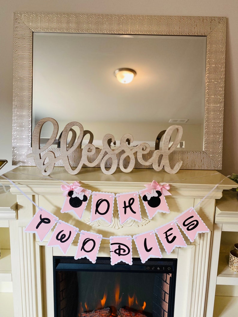 May include: Pink and white banner with the words "Oh Boy" and "Twinkle" with a Mickey Mouse head on each end. The banner is hanging above a fireplace.