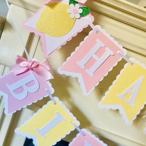 May include: A pink and yellow banner with the word "Birthday" spelled out in white letters. Each letter is on a separate flag with scalloped edges. The flags are strung together with white ribbon. There are small pink bows and a lemon with green leaves and a white flower on the banner.