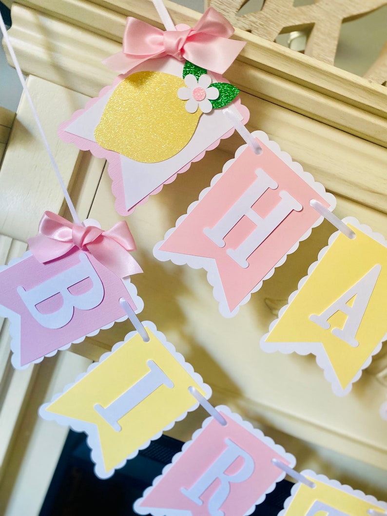 May include: A pink and yellow banner with the word "Birthday" spelled out in white letters. Each letter is on a separate flag with scalloped edges. The flags are strung together with white ribbon. There are small pink bows and a lemon with green leaves and a white flower on the banner.
