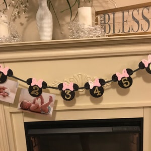 May include: A black and gold glitter banner with Minnie Mouse head shapes, pink bows, and numbers 1 through 6. The banner holds photos of a baby. The banner is strung across a fireplace mantel.