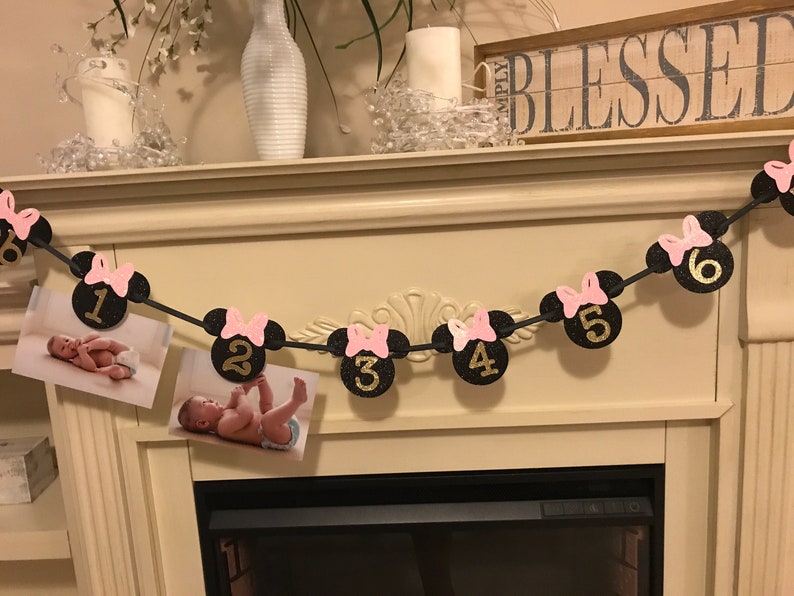 May include: A black and gold glitter banner with Minnie Mouse head shapes, pink bows, and numbers 1 through 6. The banner holds photos of a baby. The banner is strung across a fireplace mantel.