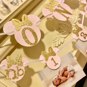May include: Pink and gold glitter Minnie Mouse themed first birthday banner with numbers 0, 1, and 2. Each number is on a pink circle with a gold glitter bow. The banner is strung together with white ribbon.