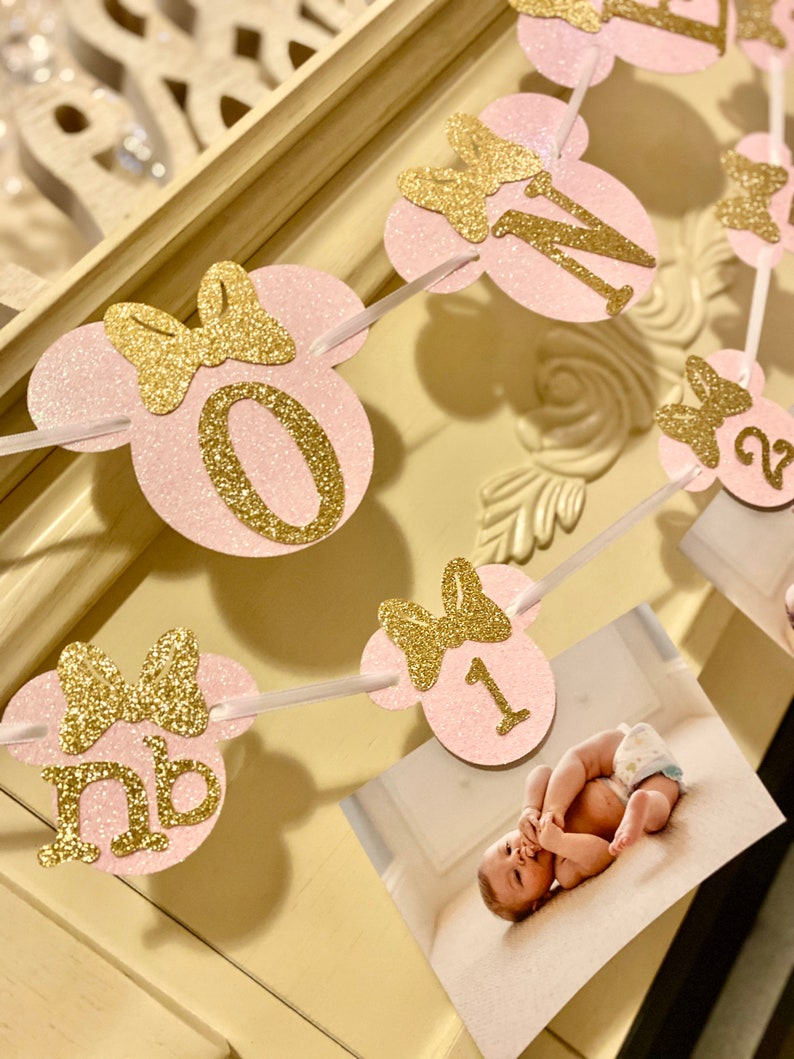 May include: Pink and gold glitter Minnie Mouse themed first birthday banner with numbers 0, 1, and 2. Each number is on a pink circle with a gold glitter bow. The banner is strung together with white ribbon.
