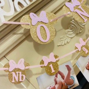 May include: Gold glitter Minnie Mouse themed first birthday banner with pink bows and numbers 0, 1, and NB. The banner is strung with pink ribbon and has photos attached.