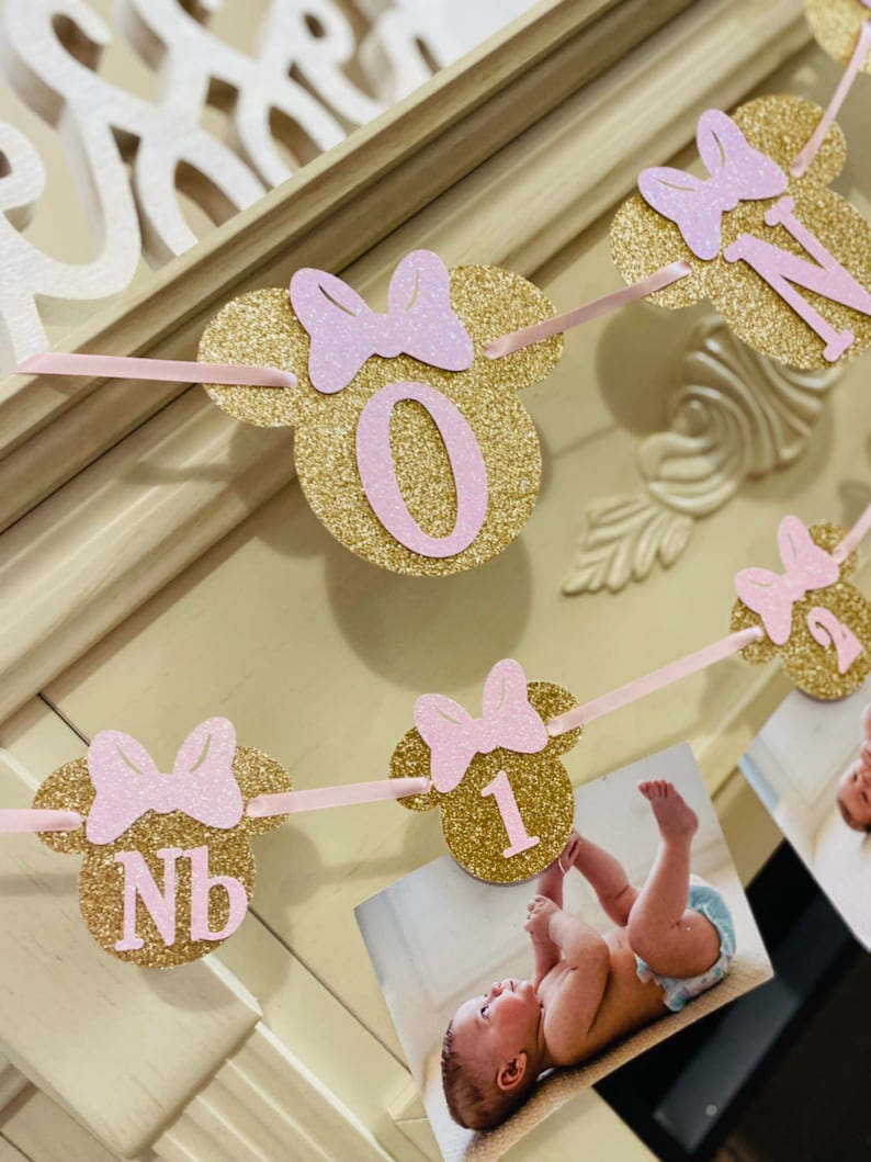 May include: Gold glitter Minnie Mouse themed first birthday banner with pink bows and numbers 0, 1, and NB. The banner is strung with pink ribbon and has photos attached.