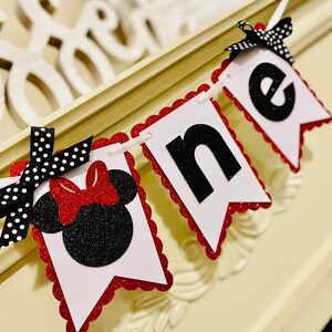 May include: A decorative banner featuring three white pennants with scalloped red glitter edges. The first pennant displays a black glitter mouse silhouette with a red glitter bow. The next two pennants feature black glitter letters spelling one.