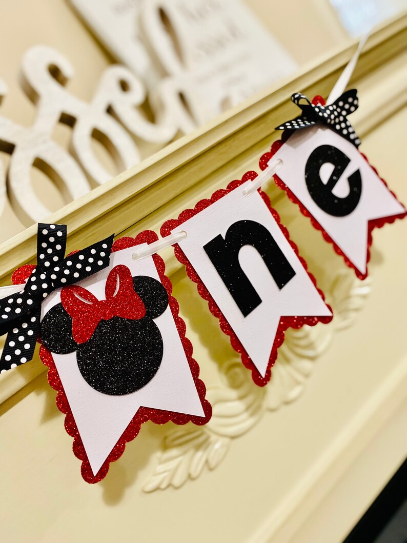 May include: A decorative banner featuring three white pennants with scalloped red glitter edges. The first pennant displays a black glitter mouse silhouette with a red glitter bow. The next two pennants feature black glitter letters spelling one.