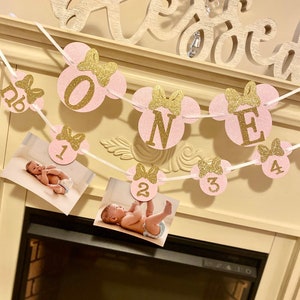 May include: Pink and gold glitter Minnie Mouse themed banner with the word "ONE" and numbers 1-4. The banner is strung with white ribbon and has photos of a baby in each section.