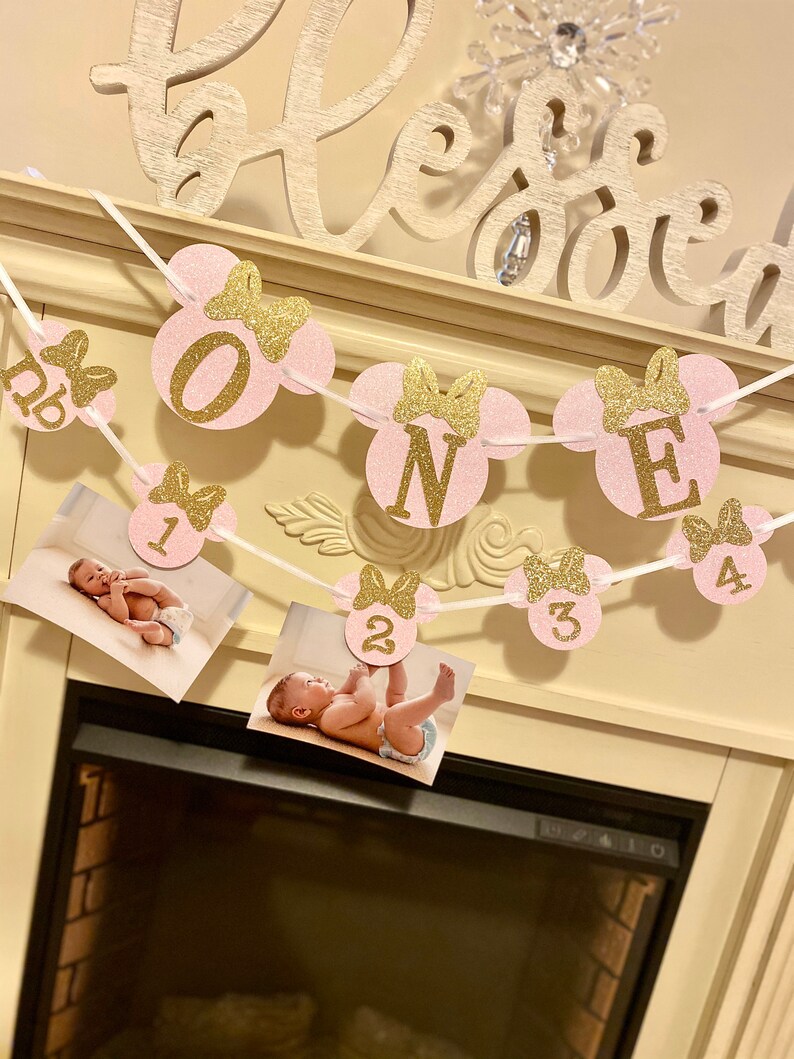May include: Pink and gold glitter Minnie Mouse themed banner with the word "ONE" and numbers 1-4. The banner is strung with white ribbon and has photos of a baby in each section.
