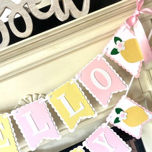 May include: A decorative banner spelling out "LILLY" in white letters on pink and yellow pennants. The banner is adorned with lemon and flower cutouts and tied with a pink ribbon. The banner is displayed on a white mantel.