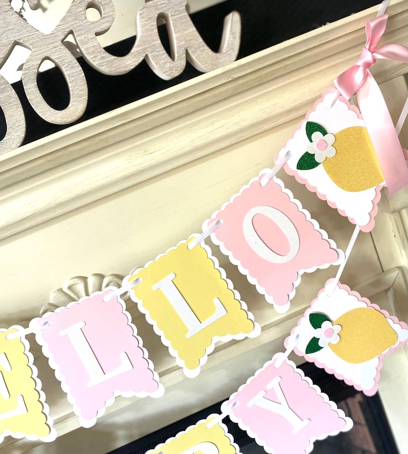 May include: A decorative banner spelling out "LILLY" in white letters on pink and yellow pennants. The banner is adorned with lemon and flower cutouts and tied with a pink ribbon. The banner is displayed on a white mantel.