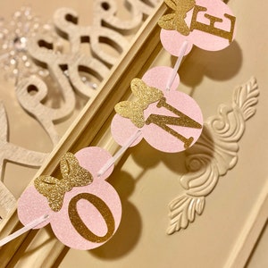 Op de afbeelding: Roze en gouden glitterbanner met de letters "ONE" in gouden glitter en een strik op elke letter. De banner is aan elkaar geregen met wit lint.
