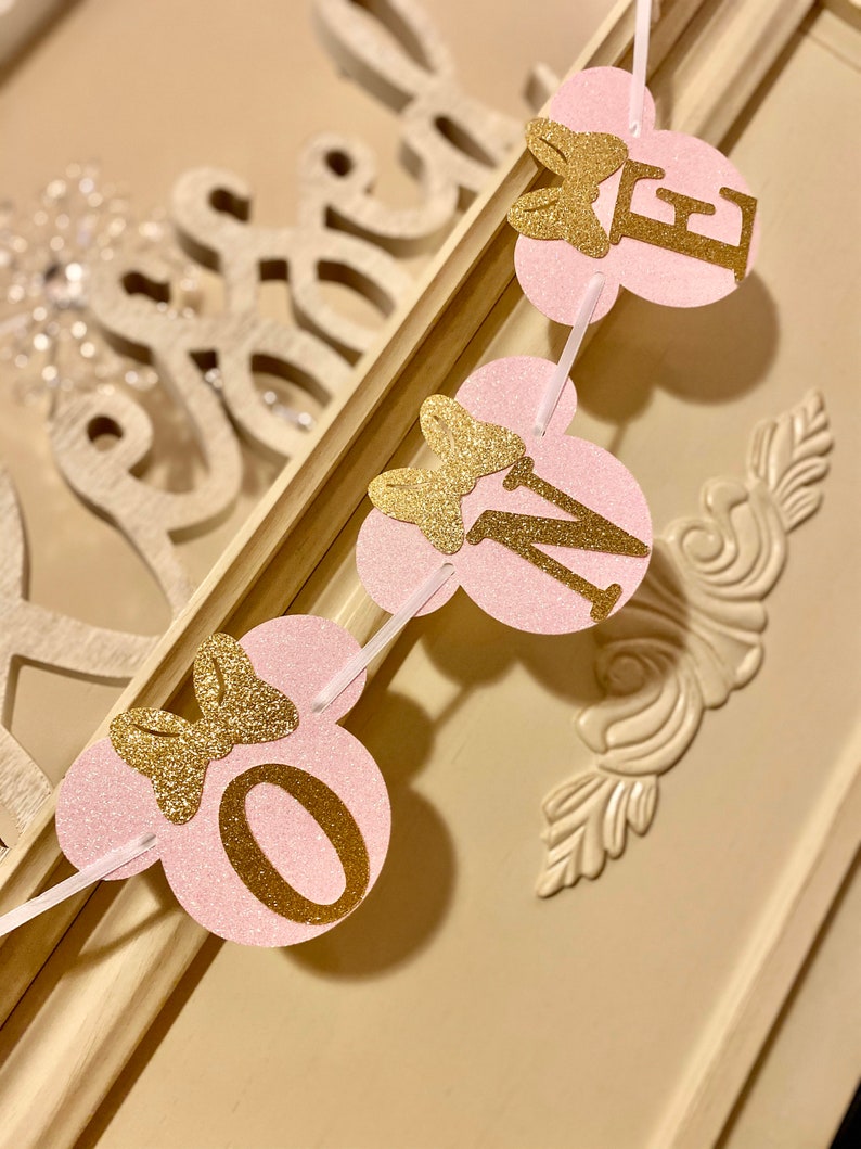 Op de afbeelding: Roze en gouden glitterbanner met de letters "ONE" in gouden glitter en een strik op elke letter. De banner is aan elkaar geregen met wit lint.