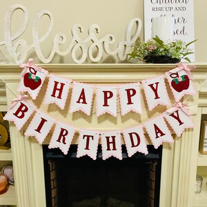 May include: Pink and white banner with red letters that reads "Happy Birthday". The banner is decorated with red apples with green leaves. The banner is hanging above a fireplace with a white wooden sign that reads "Her children rise up and call her blessed."
