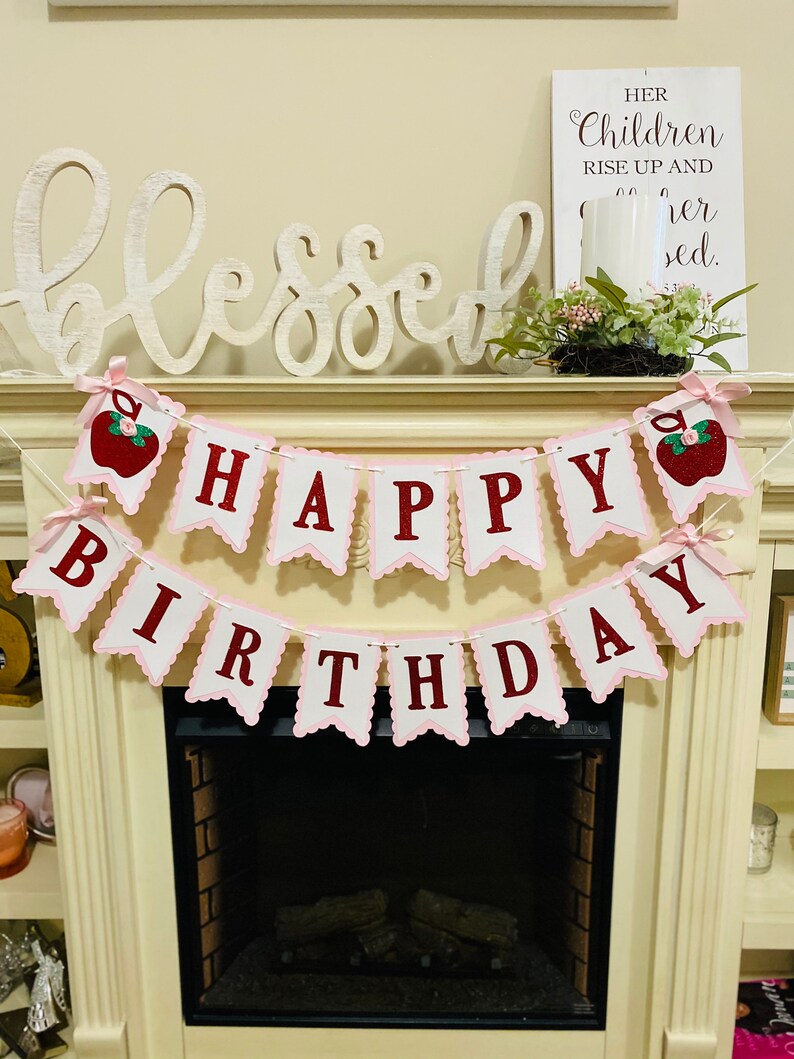 May include: Pink and white banner with red letters that reads "Happy Birthday". The banner is decorated with red apples with green leaves. The banner is hanging above a fireplace with a white wooden sign that reads "Her children rise up and call her blessed."