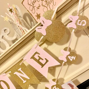 Puede incluir: Una pancarta de brillo rosa y dorado con la palabra "ONE" y orejas de Mickey Mouse con lazos. La pancarta est&aacute; decorada con los n&uacute;meros del 1 al 5.