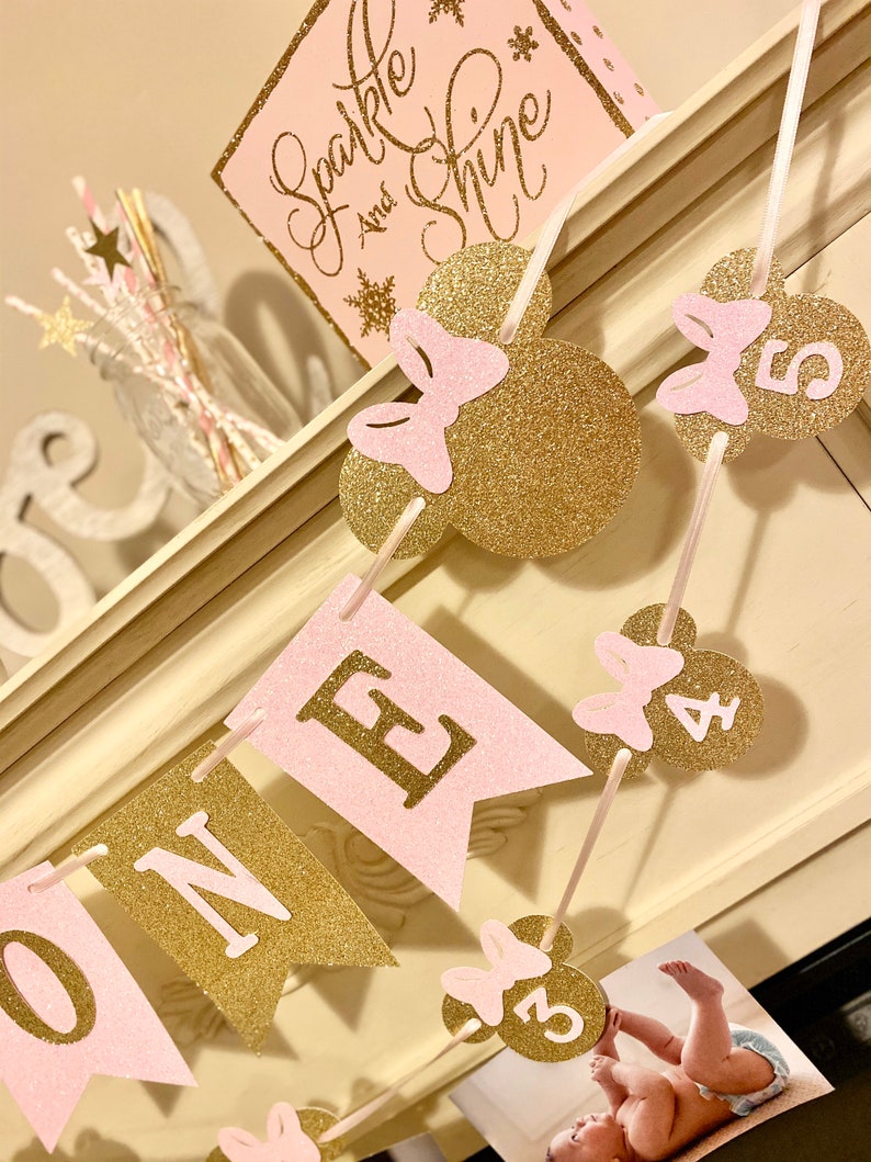 Puede incluir: Una pancarta de brillo rosa y dorado con la palabra "ONE" y orejas de Mickey Mouse con lazos. La pancarta est&aacute; decorada con los n&uacute;meros del 1 al 5.