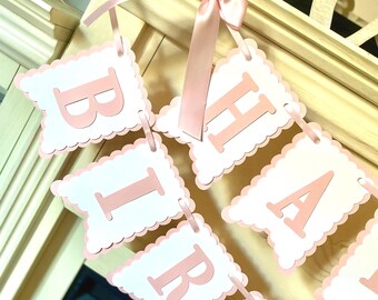 May include: A decorative banner featuring white scalloped-edge square cards with light pink lettering that spells out HAPPY BIRTHDAY. The cards are strung together with a light pink satin ribbon tied in a bow at the top, creating a festive display.