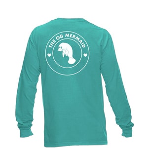Puede incluir: Una camiseta de manga larga turquesa con un gráfico circular blanco en la espalda. El gráfico presenta un manatí con el texto "THE OG MERMAID" alrededor.