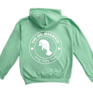 Puede incluir: Una sudadera con capucha de color verde claro con un gráfico circular blanco en la espalda. El gráfico presenta un manatí, las palabras "The OG Mermaid", dos corazones y el texto "Stay Cozy Tees".