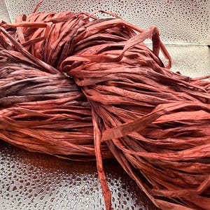 Pode incluir: Um close-up de uma bola de fita de ráfia castanho-avermelhada. A ráfia está bem enrolada, com fios individuais visíveis. A textura parece ligeiramente áspera e a cor varia de tons de vermelho claro a escuro.