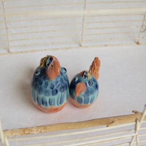 Könnte beinhalten: Zwei Keramik-Vogelfiguren in einem weißen Drahtkäfig. Die Vögel sind blau, grün und orange mit einem gefleckten Muster. Der Käfig hat eine Holzstange.