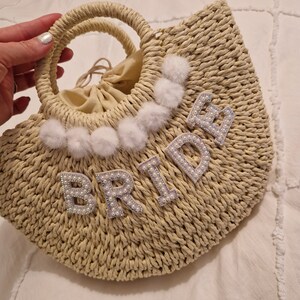 May include: A straw tote bag with a white pom pom and the word "BRIDE" spelled out in white letters with pearl accents.