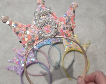 Puede incluir: Una colección de diademas con lentejuelas de colores, incluyendo una corona rosa con el número "5" adornado con perlas. Otras diademas presentan colores pastel como el morado, el azul y el amarillo. Las diademas son sostenidas por una persona.