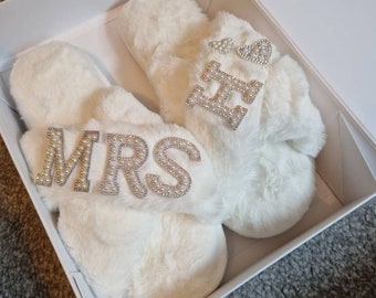 May include: A pair of white fuzzy slippers with the words "MRS" and "Mr" on them. The slippers are in a white box.