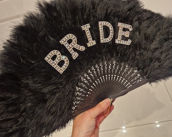 Puede incluir: Abanico de plumas negras con un adorno de "BRIDE" de strass. El abanico está cerrado y tiene un mango de madera negro.