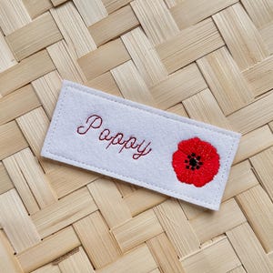 May include: A rectangular white felt patch featuring the embroidered word "Poppy" in red script, alongside a red poppy flower with a black centre. The patch is displayed on a woven bamboo background.