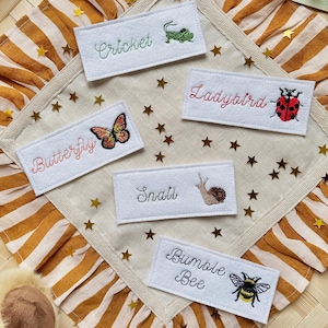 May include: Embroidered insect patches featuring a cricket, ladybird, butterfly, snail, and bumble bee. Each patch has the insect's name in script. The patches are on a cream square with a striped, ruffled border and gold star accents.