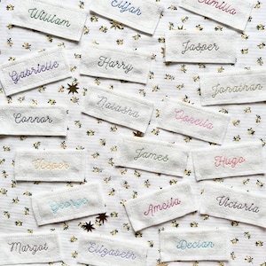 May include: A collection of white fabric labels with embroidered names in various colours, including blue, green, pink, purple, and yellow. The labels are arranged on a white background with a floral pattern.