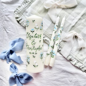 May include: Two white candles with blue and green floral designs and a larger white candle with the same design and the text "Elizabeth & Christopher 05-08-24" painted on it.
