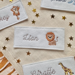 Peut inclure: Étiquettes de nom d'animaux en feutre blanc avec des détails brodés. L'étiquette en vedette indique "Lion" avec une illustration de lion assortie. D'autres étiquettes incluent un singe, une girafe et un moineau. Des confettis dorés en forme d'étoile sont dispersés autour des étiquettes.