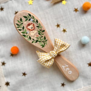 May include: Wooden baby brush featuring a hand-painted lion design framed by green leaves and white dots. A yellow and white gingham bow is tied to the handle. The name "Leo" is written in white script on the handle.