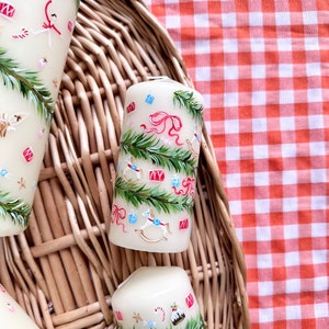 May include: Ivory-colored pillar candles with hand-painted Christmas-themed designs. The candles feature green fir branches, red bows, blue gift boxes, and toy rocking horses. The candles are arranged in a woven basket on a red and white checkered tablecloth.