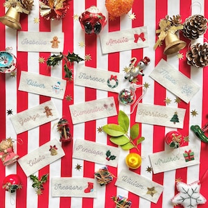 May include: A collection of embroidered name tags with Christmas decorations. Each tag displays a name in cursive, alongside embroidered festive motifs such as gingerbread men, snowflakes, and Christmas trees. The background is a red and white striped pattern.